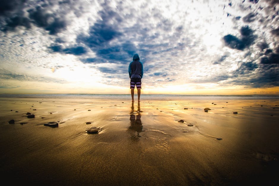 A lone figure stands on a tranquil beach during a vivid sunset, reflecting on the wet sand.